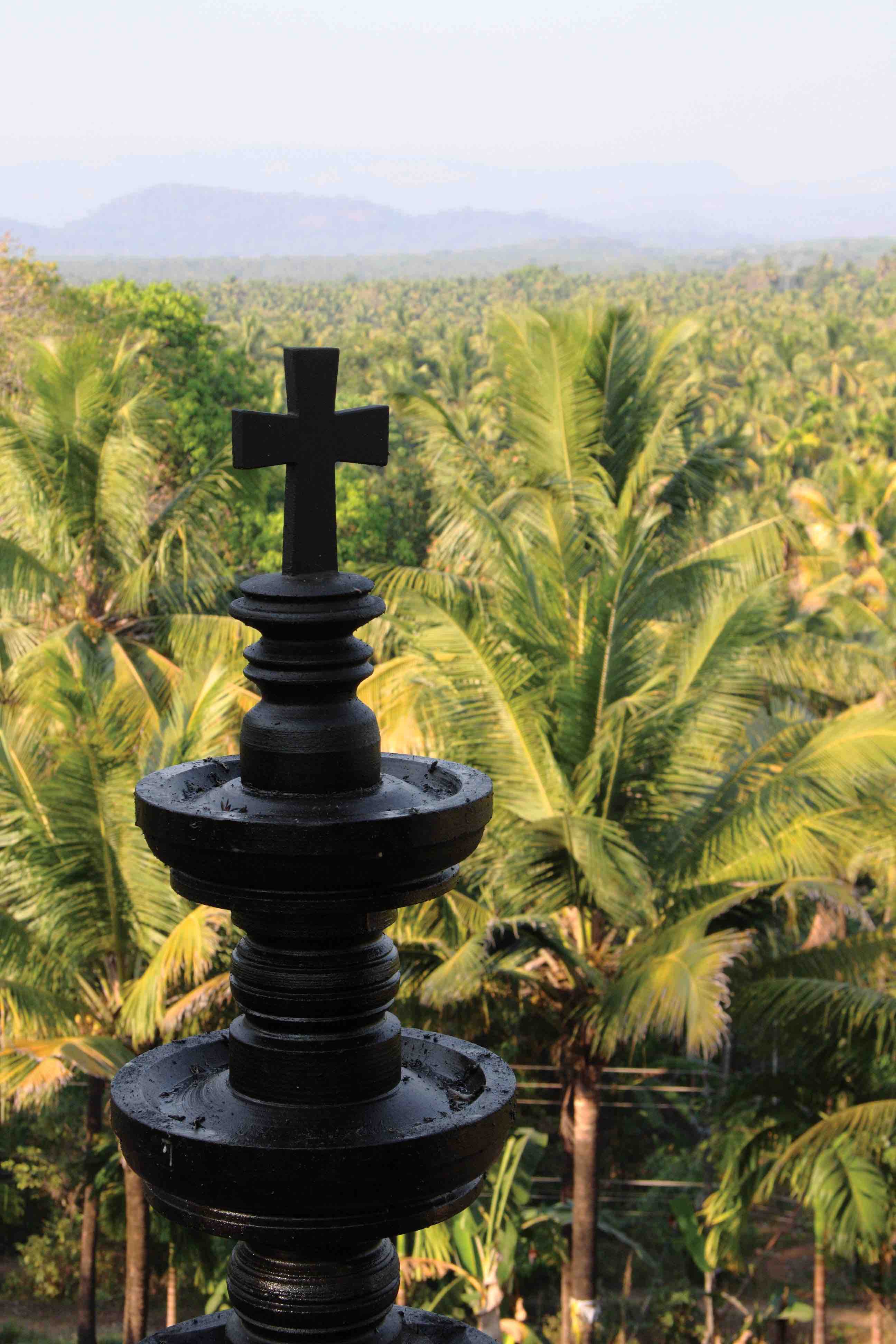 Traditional Indian stone oil lamp, topped with a cross, overlooks a coconut palm forest at the hill village of Pushpagiri